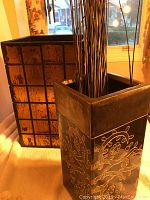 Photo of decorative wood box with gold over black grid pattern and ceramic vase with floral embossment and tall dried twigs, window background.
