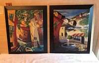 Full view of two large canvas on wood paintings with black frames showing colorful village street scenes.