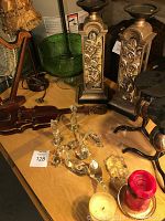 Photo showing the pair of ornate metal candleholders with floral relief on mirrored base, wooden violin without strings, glass figurines, and candles on wooden table.