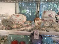 Wide view of the vintage decorative dishes with rosette floral patterns displayed on shelves, showing multiple plates and platters, some with gold trim and soft pastel floral designs.