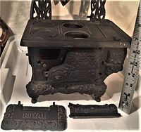 Full view of miniature cast iron Royal stove with two detached ornate parts in front, showing intricate cast details and size reference with ruler (approximately 6 inches tall).
