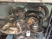 Full view of metal serving platters and other dishes arranged on metal rack shelf showing different shapes and sizes with Wilton branding visible on some handles.
