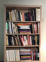 Full view of four wooden shelves filled with various cookbooks and related culinary books, some leaning and some stacked vertically.