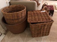Three oversized wicker baskets grouped on floor near a couch. Two are round, one is square with a lid. Lid latch on square basket needs repair.