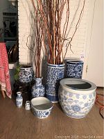 Group photo of seven blue and white Asian-style ceramic items including pots, umbrella vases, small vases, and bowl, arranged on wood floor near wall