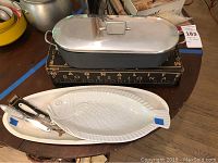 Magnalite 16-inch fish poaching pan with matching lid on original box, two white ceramic fish-shaped serving dishes below, and fish scaler on dishes.