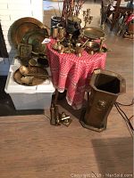 Photo showing various brass trays, cast iron honey pot with lid on a red checkered cloth table, assorted candlestick holders, ladles, and a tall hexagonal brass container on the floor.