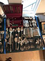 Wide view showing assorted silver plate knives, forks, spoons, serving spoons, and ladles arranged on table with velvet-lined storage cases and boxes.