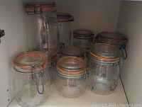 Shelf section showing a collection of large and small clear glass apothecary style jars with metal clamp lids and orange rubber seals.