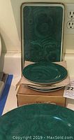 Square image showing a rectangular green swirl tray with gold trim behind four matching appetizer plates stacked on a box with a large round platter visible at the front.