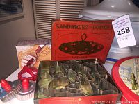 Photo showing metal animal-shaped cookie cutters stacked inside a red metal tin labeled 'Sandwiches Cookies Wonder', two plastic candy molds with red handles, and a small box with a rolling cutter.