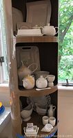 Three angled photos of brown shelf unit with white porcelain serving pieces including teapots, covered dishes, platters, egg cup, and spoon. Pieces are a mix of Arzberg (Germany), Villeroy & Boch (Luxembourg), and Apilco (France). Tape applied on some teapot spouts for protection.