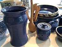 Various cobalt blue ceramic and clay serving pieces including pitchers, bowls, and small lidded containers with miniature wooden utensils, displayed on white surface.