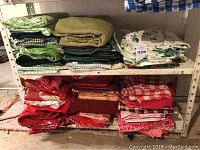 Stacked assorted green fabrics and tablecloths on top shelf and folded red fabrics and tablecloths on lower shelf of a metal rack