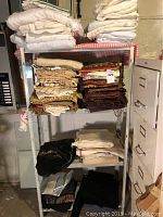 Stack of assorted folded fabrics and tablecloths on shelving, including white, brown, and patterned pieces.
