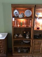Front view of the lighted wooden bookcase with five shelves showing various decorative plates, glassware, candlesticks and small ornaments on the shelves.