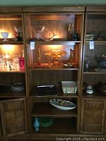 Full view of wooden lighted bookcase with glass shelves and wood paneled doors, interior lighting on, showing the front and side of the unit.