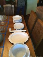 Photo of stacked white dinner plates, large platter, olive boat, and set of chairs in background