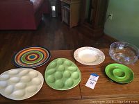 Five servingware pieces including two deviled egg plates, multicolored plate, Mikasa Pink Melody bowl, Crate and Barrel chip and dip bowl, and clear glass bowl arranged on a wooden table.