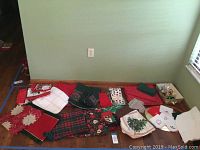 Wide shot of assorted Christmas-themed table linens including napkins, tablecloths, and placemats laid on floor against wall.