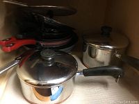 Set of stainless steel pots and pans including a large stock pot and smaller pots with black handles and lids, along with three non-stick pans with red handles stacked together.