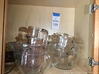 Clear glass measuring cups in assorted sizes stacked inside a cabinet, visible measurement markings in red and raised glass, all with handles.