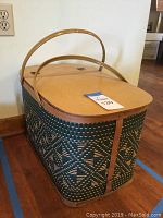 Closed woven picnic basket with wooden lid and handles in natural light against a wall.