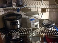 Photo of three kitchen appliances on a wire shelf including a Crock Pot slow cooker with lid, plastic steamer with spout, and waffle baker.