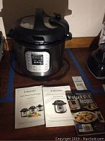 Electric pressure cooker on wooden floor with power cord with user manuals and recipe booklet in front of it