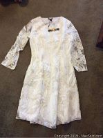 White ladies dress with floral leaf pattern and 3/4 sleeves laid flat on carpeted floor.