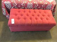 Full view of rectangular red upholstered storage bench with button tufted top, placed on carpet in front of colorful fabric background.