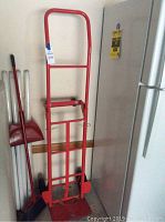 Full view of red steel dolly hand truck standing upright next to a wall with broom and dustpan nearby.