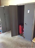 Photo showing two tall grey metal storage cabinets with doors partially open, revealing interior. One cabinet has a red suitcase inside.