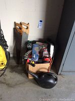 Wide shot of the box with two hand saws leaning against the wall and a black plastic watering can in front. Several tools and items packed inside the box including a flashlight.