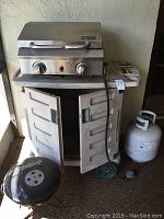 Front and side view of Sportsman Series outdoor gas grill on plastic storage cart next to propane tank and charcoal grill lid