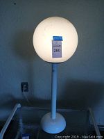 Photo of white outdoor floor lamp with round globe shade, lit on, against blue wall, showing base and power cord.
