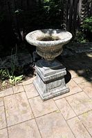 Concrete urn planter mounted on square pedestal base outdoors on paved surface, showing full lot with natural lighting and shadow.