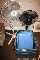 Photo showing two pedestal fans by Super, one white and one black, alongside three plastic tote bins with lids stacked in front.