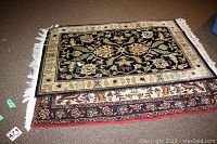 Two wool area rugs stacked on brown carpet floor, showing floral vine patterns and fringes on short sides.