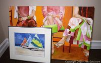 Framed watercolor painting with three sailboats and colorful sails leaning against a large canvas painting of seated women.