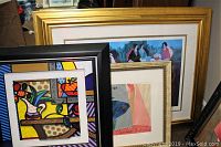 Three framed art pieces leaning against a wall: black-framed abstract, gold-framed Tarkay print, and glass art in pale frame