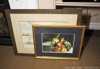 Two framed artworks side-by-side on floor, one with colorful vegetable still life, one with a tree and landscape.