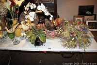 View of entire lot including glass vases with faux fruit, wooden bird statues, faux orchids, faux floral stems, and wreaths on table.