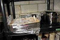 Three roasting pans placed on a black shelf wrapped in plastic. Two are stainless steel pans with racks, one lidded roaster on the right side.