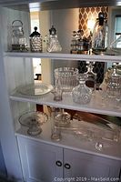 Three shelves filled with clear glassware, including vases, decanters, lidded jars with metal tops, cake pedestal, and small glass basket on white shelves.