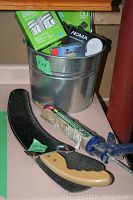 Photo showing metal bucket filled with various boxed lightbulbs, caulking gun, saw, and hammer.