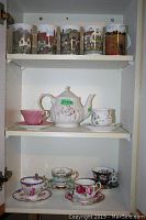 Full view of shelves showing teapot with pink floral design and six assorted tea cups with saucers below, Dunoon 'Country Cottages' mugs on top shelf