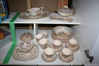 China set displayed on shelf with visible tea cups, saucers, dinner plates, platter and gravy boat showing floral and decorative brown border pattern.