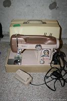Front view of White sewing machine with beige and metallic brown finish, attached foot pedal and hard case behind.