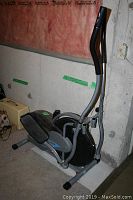 Side view of OrbiTrek Platinum elliptical trainer against basement wall, showing handles, foot pedals, and base with manual resistance knob.